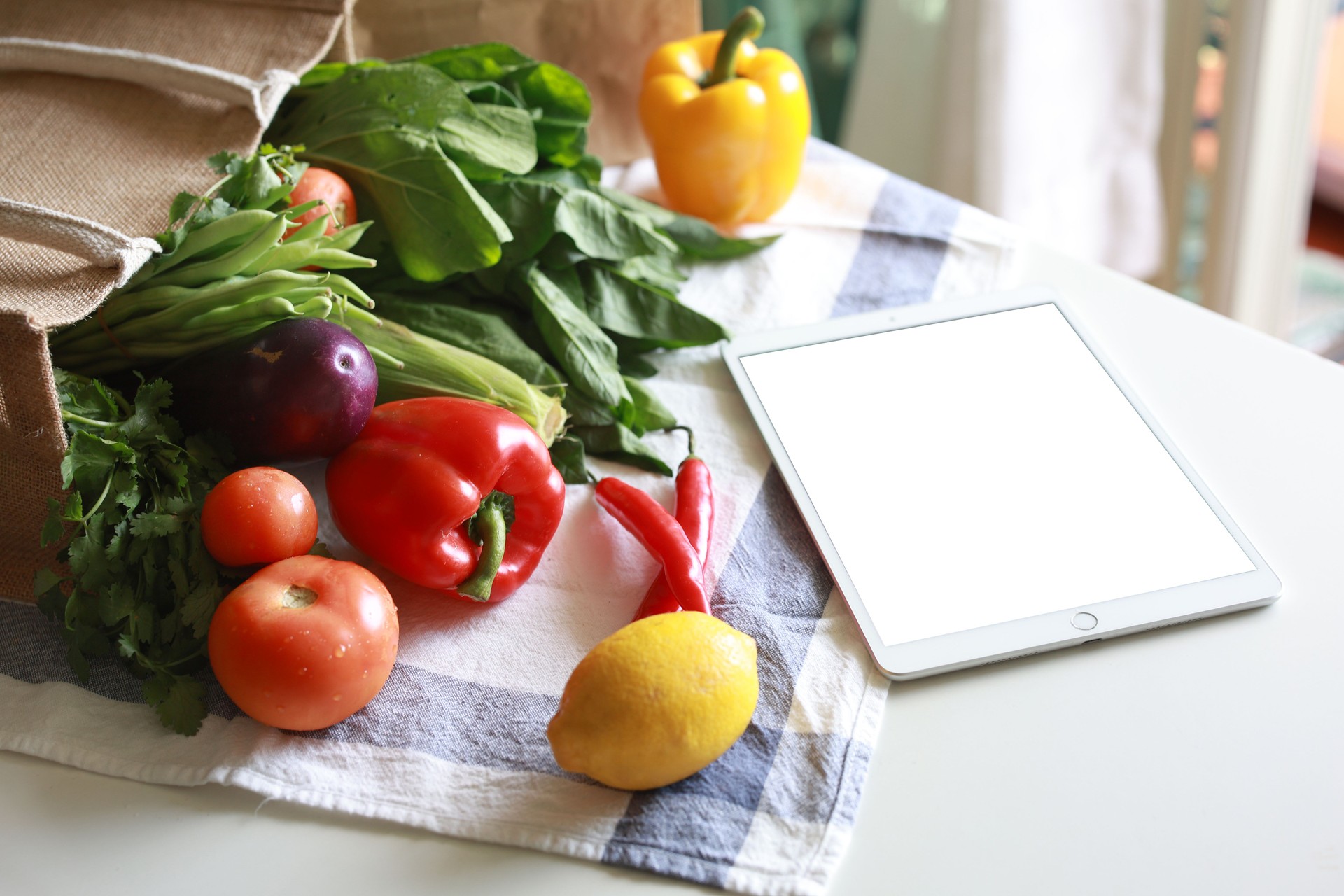 Une tablette numérique à écran blanc et un sac à provisions réutilisable écologique rempli de légumes frais multicolores et de produits d’épicerie sur le comptoir de la cuisine à la maison.
